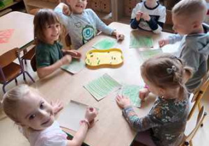 Grupa dzieci siedzi przy stoliku i koloruje przy użyciu zielonych pasteli olejnych powierzchnię kartonu
