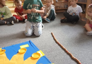 Chłopiec nawleka kolejny koralik na "łańcuch życia", w tle widać siedzi siedzące w urodzinowym kręgu na dywanie