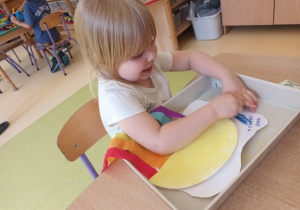 Dziewczynka siedzi przy stoliku i koloruje na swojej pracy plastycznej chmurę niebieską pastelą