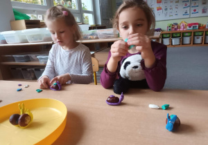 Dziewczynki siedzą przy stoliku i lepią ślimaki z plasteliny, przed nimi na stoliku leżą ulepione wcześniej ślimaki