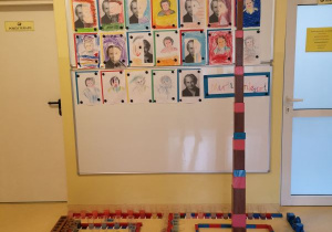 Zdjęcie przedstawia wystawę prac dzieci, na której znajdują się portrety Marii Montessori namalowane przez dzieci, różowa wieża, brązowe schody, czerwono niebieskie sztangi, czerwone sztangi, kubeczki z zafarbowaną na różne kolory wodą, tabliczki barwne, kolorowe cylindry, cylindry do osadzania, dwa napisy Maria Montessori ułożone z ruchomego alfabetu