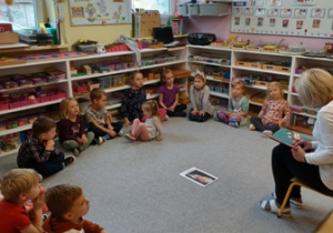 dzieci siedzą na dywanie w kręgu, na środku leży portret Marii Montessori. Na krzesełku przed dziećmi siedzi pani.