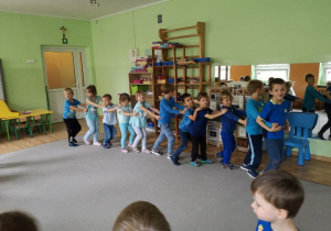 Dzieci ubrane w niebieskie stroje podczas zabawy ruchowej, ustawione w pociąg idą jedno za drugim