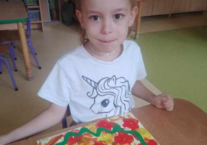 Dziewczynka siedzi przy stoliku i pozuje do zdjęcia, na stoliku widać pracę plastyczną zrobioną z okazji "Dnia Spadającego Liścia"