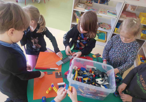 Dzieci stoją przy stole i budują z klocków znajdujących się w dużym pudełku