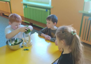 Dzieci siedzą przy stoliku, chłopiec wrzuca liście szpinaku do dzbanka