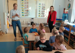 Dzieci siedzą [przy stolikach i słuchają prowadzących warsztaty kulinarne