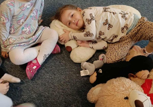 Dziewczynka siedzi na dywanie, druga dziewczynka leży wtulona w swojego misia