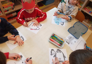 Dzieci siedzą przy stole i kolorują obrazki z pingwinami