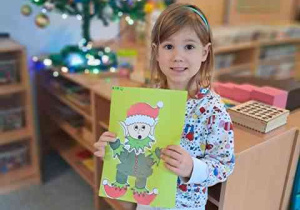 Dziewczynka na tle choinki pozuje do zdjęcia z kartą pracy pt. "Pomocnik Świętego Mikołaja - Elf"