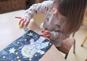 Dziewczynka posypuje swoją pracę plastyczną pt. "Astronauta w galaktyce" brokatem