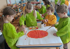 Dzieci stoją dookoła stolika i malują farbami duży karton w grupie kilkuosobowej