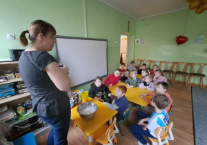 Dzieci siedzą przy stoliczkach i słuchają prowadzącej warsztat