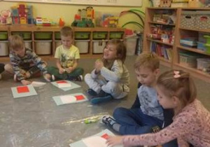 Dzieci siedzą na dywanie i wykonują pracę plastyczną - wyklejają sylwetę Mikołaja zgodnie z przedstawioną instrukcją z elementów kolorowego papieru