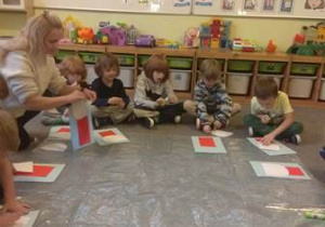 Dzieci siedzą w kręgu na dywanie i wykonują na folii pracę plastyczną pt. "Mikołaj"
