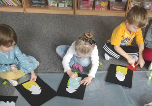 Dzieci siedzą na dywanie i przyklejają na kartkę wizerunek "Skrzata"