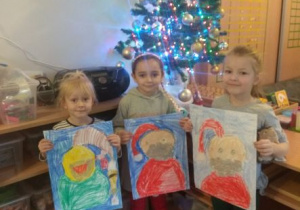 Dziewczynki stoją przy choince i prezentują narysowanego Świętego Mikołaja.