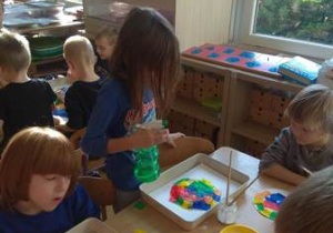 Dzieci przy stoliku w czasie eksperymentowania z barwami wykorzystują kawałki kolorowej bibuły oraz wodę