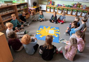 Dzieci siedzą na dywanie w kręgu. Na środku dywanu leży urodzinowe słoneczko, na którym dzieci układają koraliki symbolizujące wszystkie miesiące życia Kacperka
