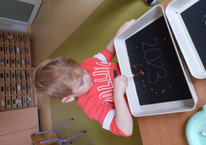 Chłopiec odbija patyczek umoczony w pomarańczowej farbie na swojej pracy plastycznej