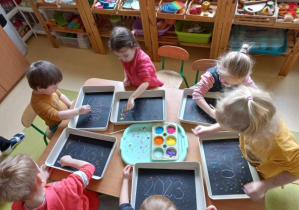 Dzieci przy stoliku stemplują wacikami kolorowe kropki na czarnym papierze