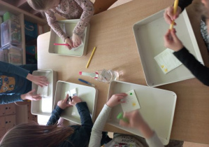 Dzieci przy stoliku rysują mazakami po chusteczce złożonej w kwadrat