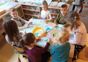 Dzieci przy stoliku przyklejają papierowe trójkąty i uzupełniają elementy drzewa
