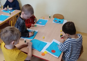 Dzieci siedzą przy stoliku i przyklejają niebieskie tło do białej kartki