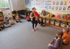 Dzieci siedzą na dywanie w czasie ćwiczenia ciszy, Nela stoi na środku i trzyma w dłoniach serduszka od dzieci z innego przedszkola