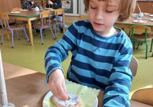 Zdjęcie prezentuje chłopca, który ozdabia swoją prace plastyczną przy użyciu kolorowej soli do kąpieli