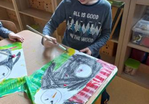 Chłopiec trzyma w dłoni pędzel i maluje tło swojej pracy plastycznej, na której widać pingwina