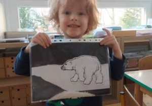Chłopiec trzyma w dłoniach wykonaną przez siebie pracę plastyczną przedstawiającą niedźwiedzia polanego na ciemnym tle