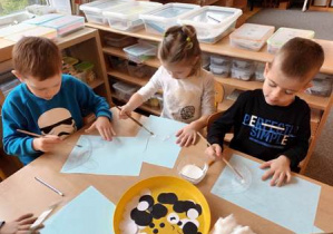 Dziewczynka i dwóch chłopców siedzą przy stoliku i rozprowadzają klej przy użyciu pędzli na narysowanej przez siebie sylwecie niedźwiedzia polarnego