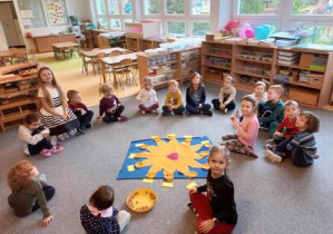 Dzieci siedzą na dywanie w kręgu, na środku leży "Urodzinowe Słoneczko", na którym dzieci układają koraliki symbolizujące wszystkie miesiące życia Amelki