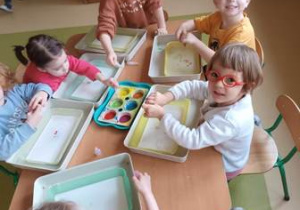 Dzieci młodsze siedzą przy stoliku i tworzą kompozycję malując farbami na mleku