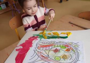 Dziewczynka siedzi przy stoliku i maluje tło swojego obrazu farbami