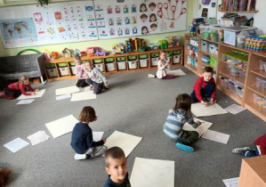 Dzieci na dywanie grają w grę "Wylosuj twarz" zainspirowaną twórczością Picasso i rysują wyniki na dużym arkuszu