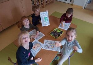 Dzieci młodsze siedzą przy stoliku i kolorują przy użyciu pasteli olejnych szablony twarzy charakterystycznych dla twórczości Picasso