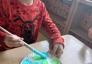 Chłopiec przy stoliku wykonuje prace plastyczną i maluje powierzchnię papierowego talerzyka rosnąca farbą