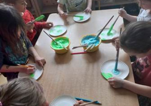 Dzieci siedzą przy stoliku i malują rosnącą farbą w kolorze zielonym oraz niebieskim talerzyk papierowy