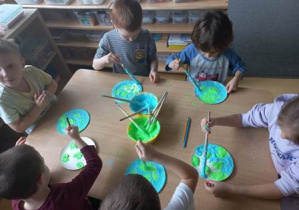 Dzieci siedzą przy stoliku i malują na papierowym talerzyku Ziemię z wykorzystaniem rosnącej farby w kolorze niebieskim oraz zielonym