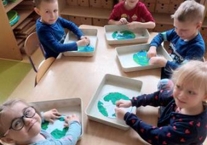 Dzieci siedzą przy stoliku i rozprowadzają paluszkami plastelinę w kolorze zielonym oraz niebieskim na szablonie Ziemi