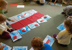 Dzieci siedzą na dywanie i tworzą prace plastyczne z flagą Polski