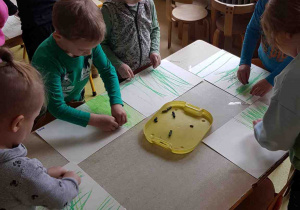 Dzieci stoją wokół stolików i rysują trawę na kartkach papieru