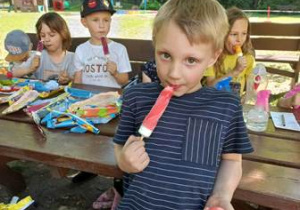 Maciek je lody, w tle widać inne dzieci siedzące na ławce w ogrodzie przedszkolnym i jedzące lody