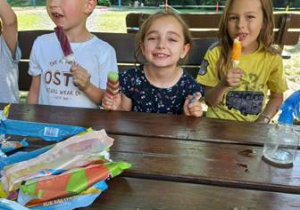 Dzieci siedzą przy stoliku w ogrodzie przedszkolnym i jedzą lody