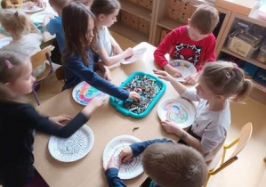 Dzieci siedzą przy stoliku i ozdabiają przy użyciu pasteli olejnych talerzyk papierowy tworząc muszlę ślimaka