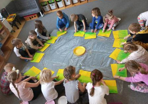 Dzieci w czasie wykonywania pracy plastycznej siedzą na dywanie i malują pastelami trawę na żółtym arkuszu papieru