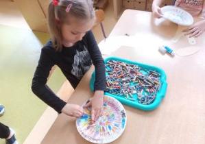 Dziewczyna siedzi przy stoliku i maluje pastelami olejnymi papierowy talerzyk