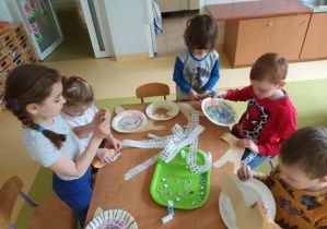 Dzieci w czasie wykonywania pracy plastycznej doklejają elementy kolorowego papieru do pomalowanego wcześniej papierowego talerzyka tworząc postać ślimaka